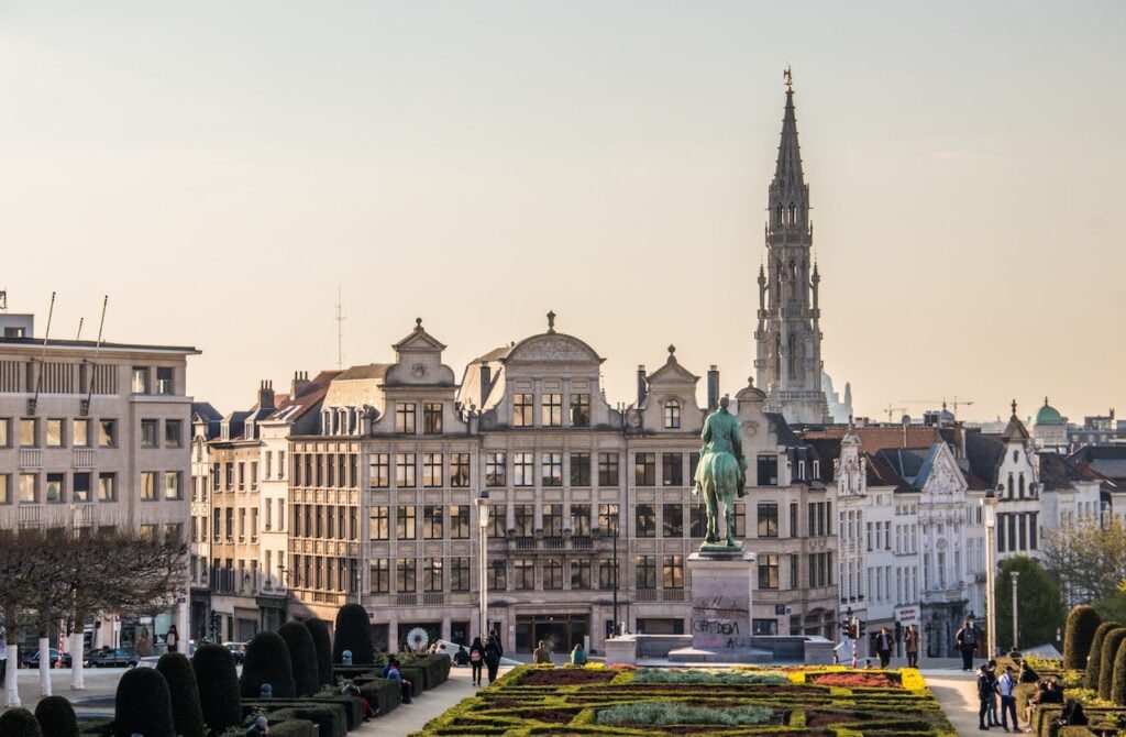 Vue panoramique d'une place de la ville de Bruxelles, en Belgique, avec ses bâtiments historiques. Au premier plan, on peut voir un jardin bien entretenu et une statue d'un personnage à cheval, tandis qu'à l'arrière-plan, la haute flèche de l'hôtel de ville s'élève au-dessus des toits.