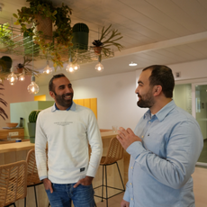 Deux hommes discutent et sourient dans un bureau moderne avec des sièges de style bar, des plantes en pot et des ampoules suspendues en arrière-plan. L'un des hommes porte un pull blanc et l'autre une chemise bleu clair.