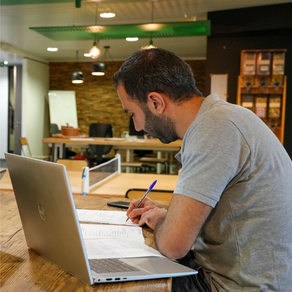 Un homme aux cheveux noirs courts et à la barbe vêtu d'un polo gris est assis à une table en bois, écrivant dans un cahier avec un stylo bleu. Un ordinateur portable ouvert est devant lui. L'arrière-plan présente un bureau moderne avec des étagères et un tableau à feuilles mobiles.