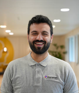 Un homme souriant aux cheveux noirs et à la barbe se tient dans une pièce bien éclairée, vêtu d'un polo gris avec un logo sur lequel on peut lire « iterates » et « IT services & consulting ». L'arrière-plan est légèrement flou avec un mur jaune et blanc et quelques plantes d'intérieur vertes.