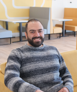 Un homme barbu au sourire amical est assis sur un canapé jaune. Il porte un pull rayé gris et noir. À l'arrière-plan, des sièges modernes et confortables dans un environnement décontracté et bien éclairé.