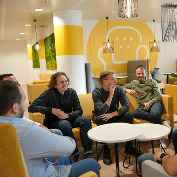 Un groupe de sept personnes sont assises autour de deux tables basses blanches dans un salon de bureau confortable et moderne aux murs jaunes et blancs. Elles discutent en toute émotion, sourient et rient, assises sur des canapés et des chaises jaunes.