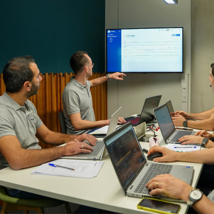 Quatre personnes sont assises autour d'une table avec des ordinateurs portables et participent à une discussion collaborative. Une personne pointe du doigt un écran sur le mur affichant un document ou une présentation. Des blocs-notes, des téléphones et divers autres objets sont répartis sur la table.