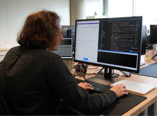 Une personne aux cheveux longs est assise à un bureau et travaille sur un ordinateur doté de deux écrans. Un écran affiche du code, tandis que l'autre affiche un e-mail ou un document. Ils utilisent un clavier et une souris, et des fournitures de bureau supplémentaires et un ordinateur portable sont posés sur le bureau.