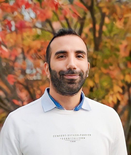 Un homme aux cheveux noirs courts et à la barbe sourit devant un arrière-plan de feuilles d'automne colorées. Il porte une chemise blanche avec un col bleu. Les arbres flous présentent une gamme de feuillages orange, rouges et jaunes.