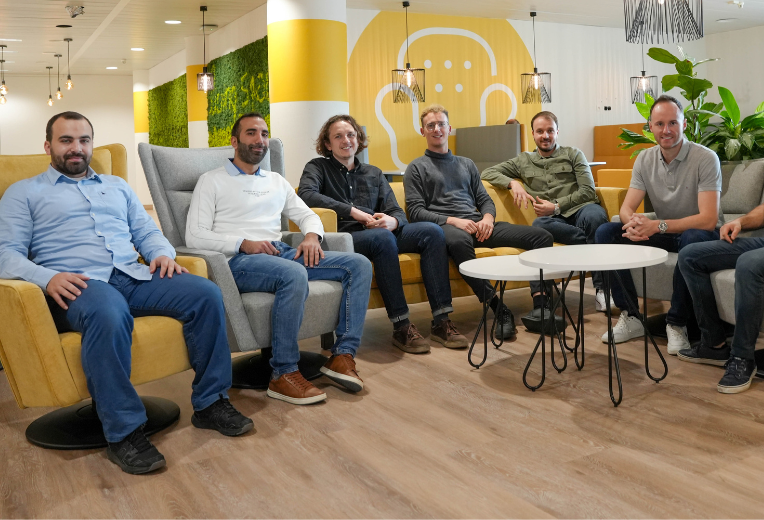 Un groupe de sept personnes est assis dans un salon de bureau moderne, quatre d'entre elles dans des fauteuils et trois sur un canapé. Ils sont souriants et décontractés. L'arrière-plan présente des murs jaunes et blancs, des plantes vertes et une décoration élégante.